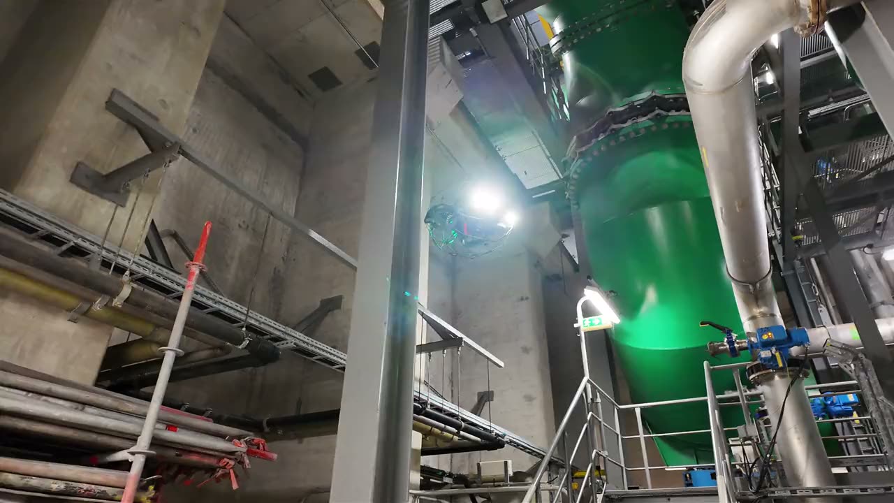 Die Elios 3 im Schutzkäfig fliegt mit eingeschaltetem Licht vor einer großen Rohrleitung in einer Industrieanlage