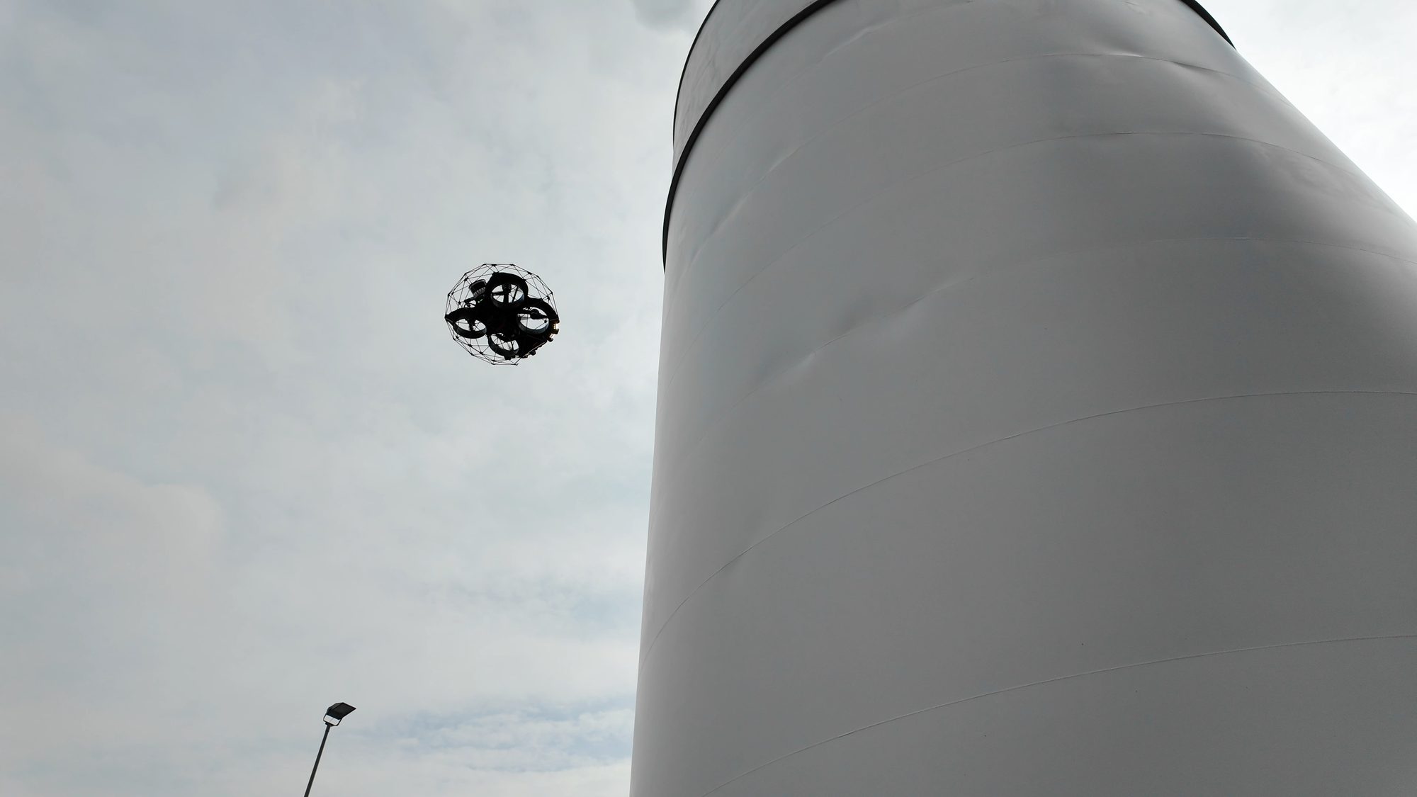 Inspektionsdrohne im Schutzkäfig fliegt neben einem grauen Stahltank