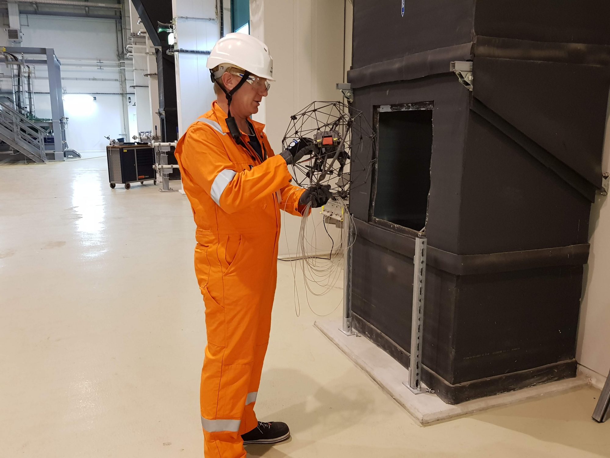 Techniker in orangem Schutzanzug hält eine Inspektionsdrohne im Schutzkäfig an eine Gehäuseöffnung
