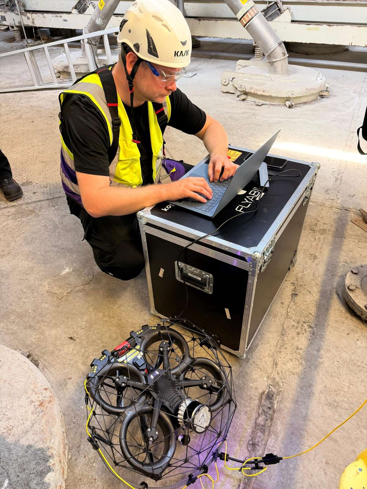 Pilot mit Helm kniet am Transportkoffer und arbeitet am Laptop, davor die Drohne im Käfig