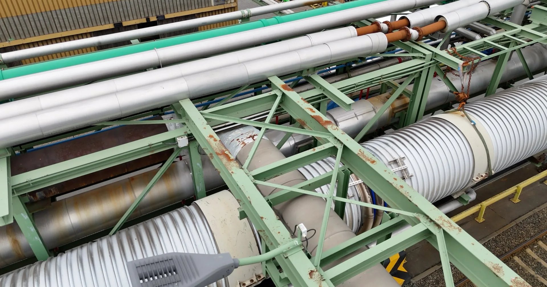 Rohrbrücke von oben aus dem Drohnenflug: grüne Stahlträger mit Rostflecken, darunter gedämmte Rohrleitungen mit Blechmantel und Kompensatoren, daneben Gleise