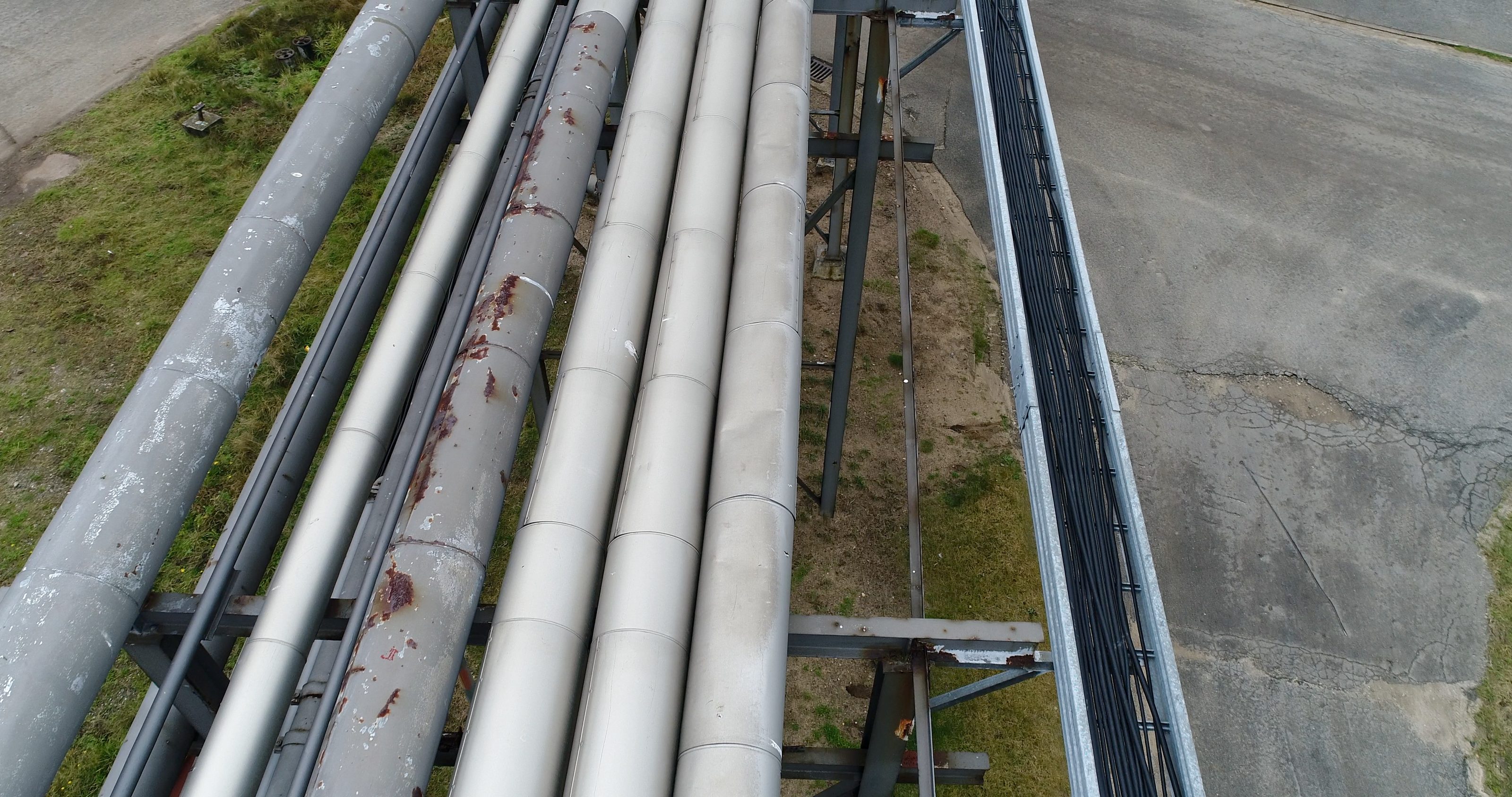 Rohrtrasse längs von oben: Rost am Mantel einer gedämmten Leitung und an Querträgern der Stützkonstruktion, rechts die Kabeltrasse über Asphalt
