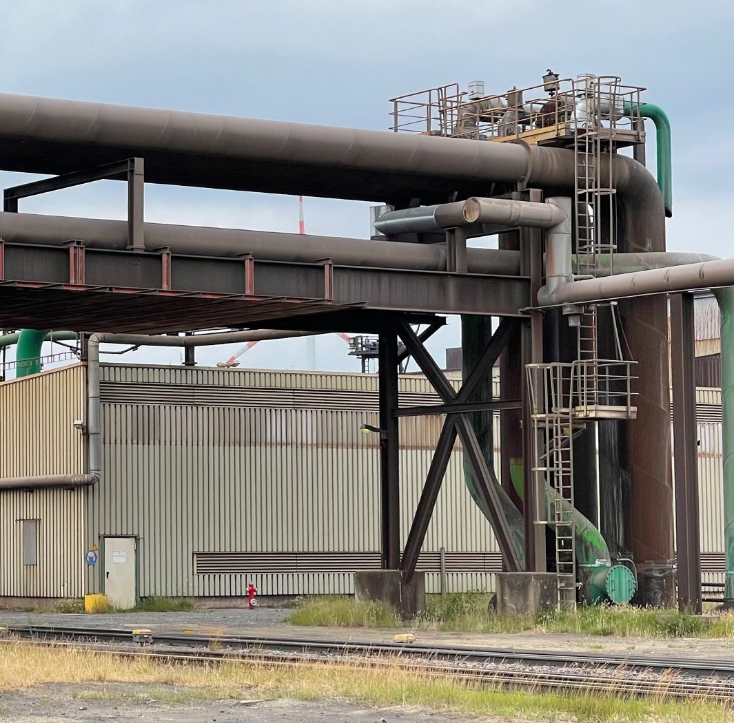Stählerne Rohrbrücke mit mehreren Rohrleitungen, Stützen und Steigleitern über Werksgleisen, dahinter eine Industriehalle
