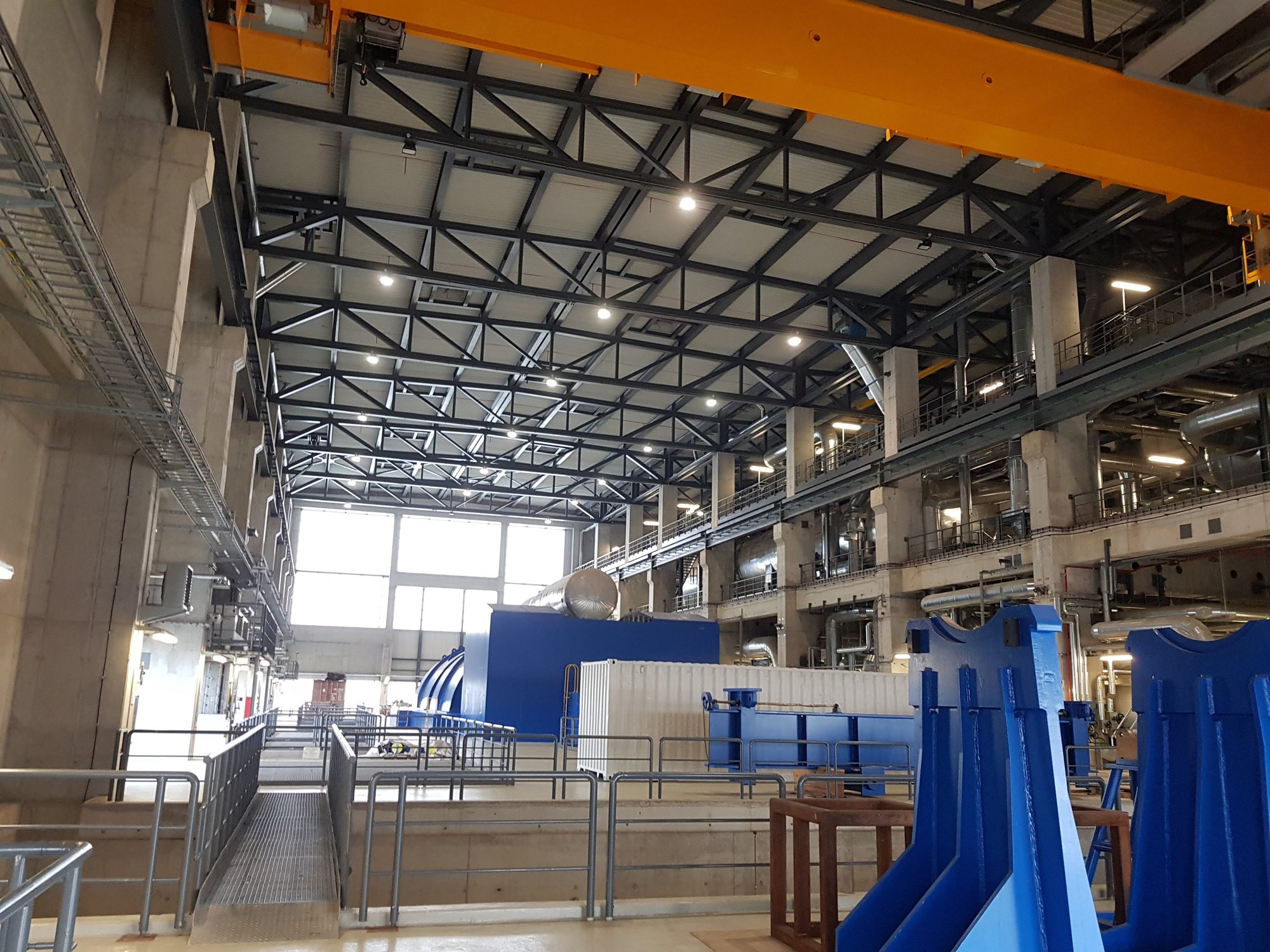 Maschinenhalle eines Kraftwerks: oben ein gelber Brückenkran und das Dachtragwerk aus Stahlfachwerk, unten ein Maschinensatz in blauem Gehäuse, rechts Bühnen mit gedämmten Rohrleitungen
