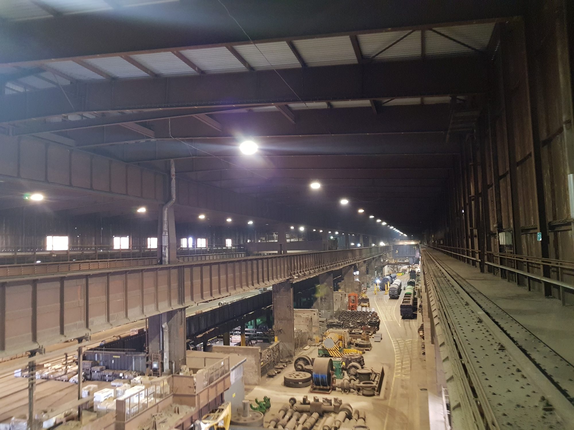 Lange Stahlwerkshalle von oben: links ein genieteter Kranbahnträger auf Stützen, rechts Kranschiene und Laufsteg, unten Lagerflächen und Gleise mit Waggons