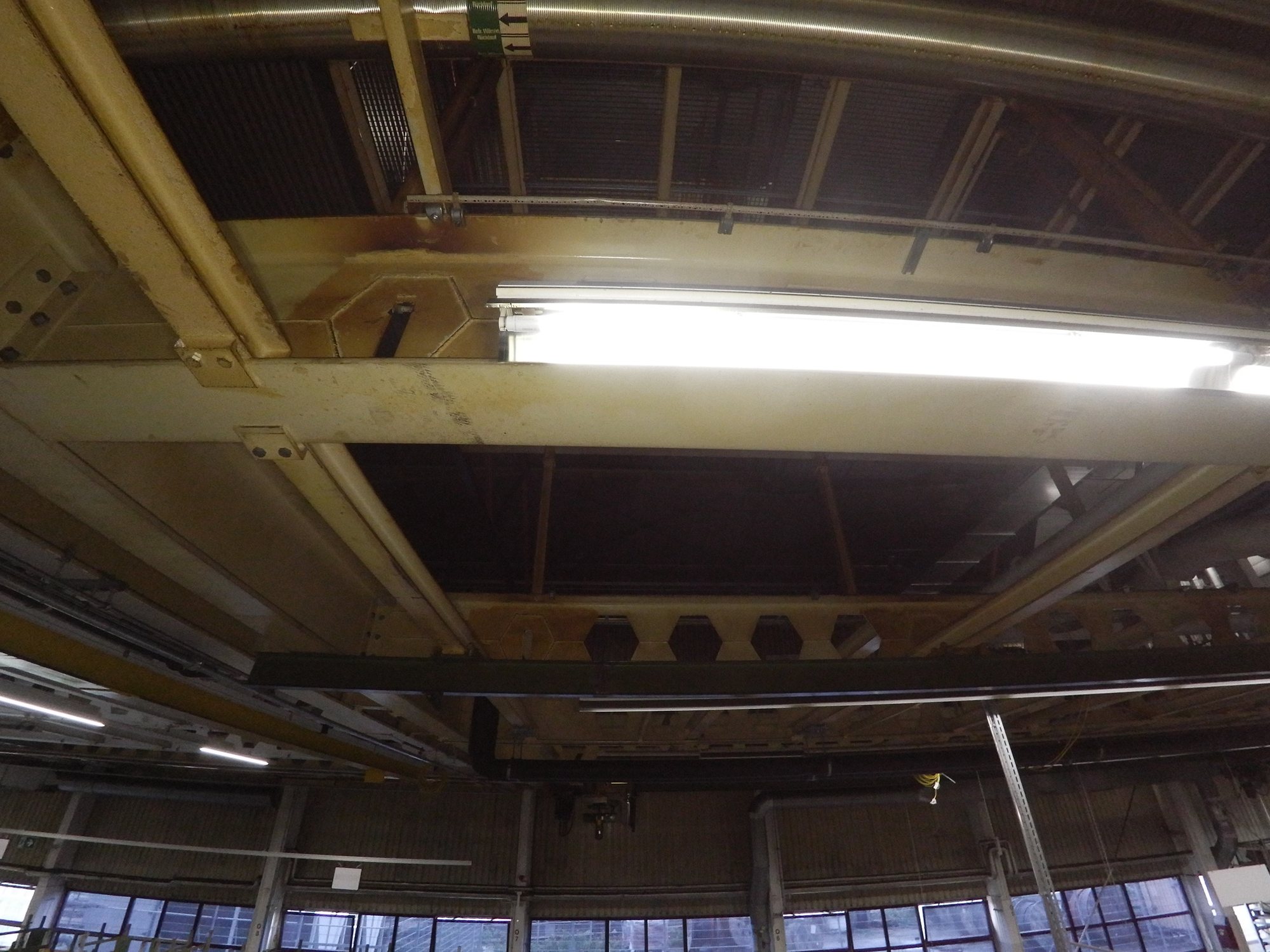 Blick von unten auf das Stahltragwerk einer Hallendecke mit Trägern, Knotenblechen, Leuchte und Lüftungsrohr; an Trägern und Knotenblechen braune Verfärbungen