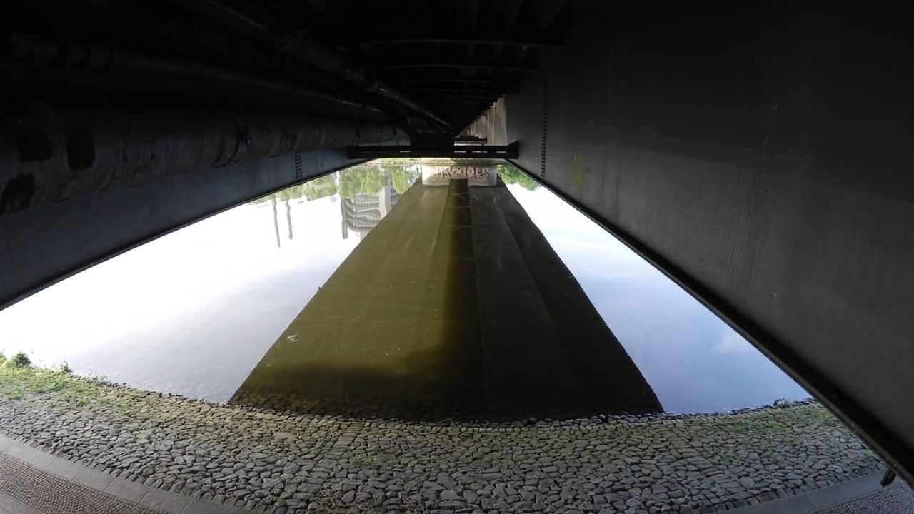 Blick vom Ufer unter eine Straßenbrücke, das Wasser spiegelt den Himmel, darüber der Überbau