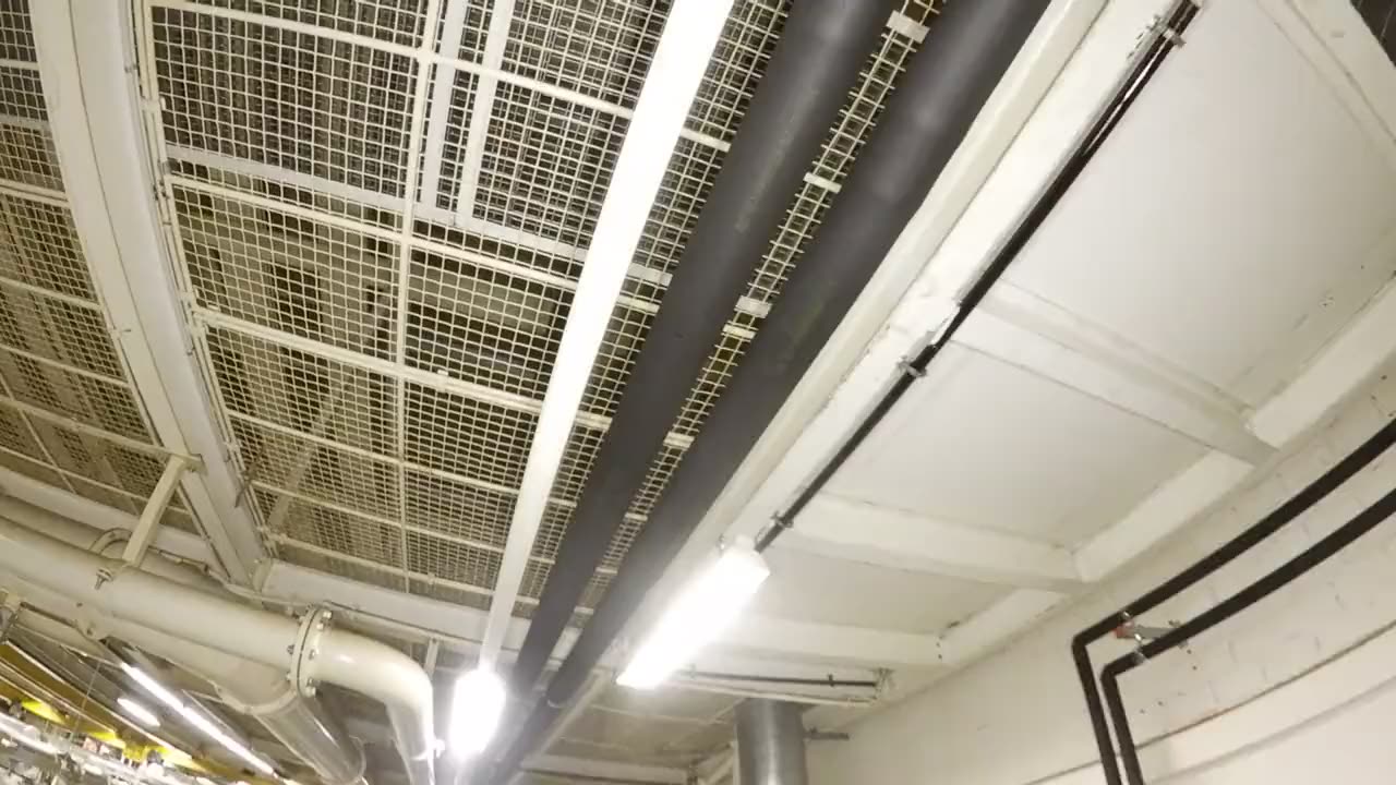 Drohnenflug unter einer Hallendecke mit Gitterdecke, Rohrleitungen und Leuchtenband