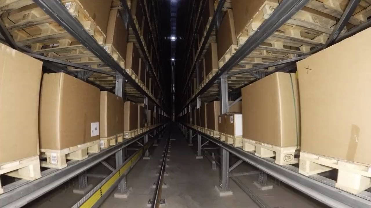 Drohnenflug in der Mitte einer schmalen Regalgasse, links und rechts Paletten mit Kartons in vielen Ebenen