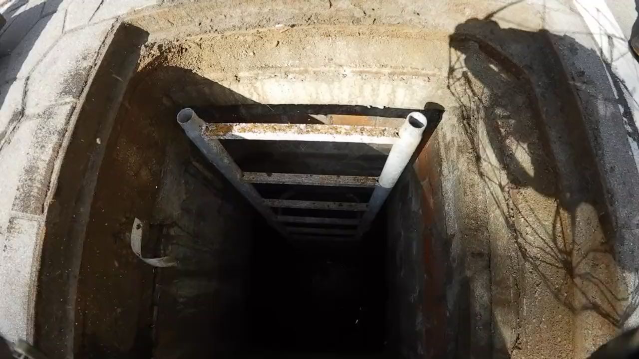 Blick von oben in einen engen Einstiegsschacht mit Steigleiter, ringsum befestigte Fläche, unten das dunkle Becken